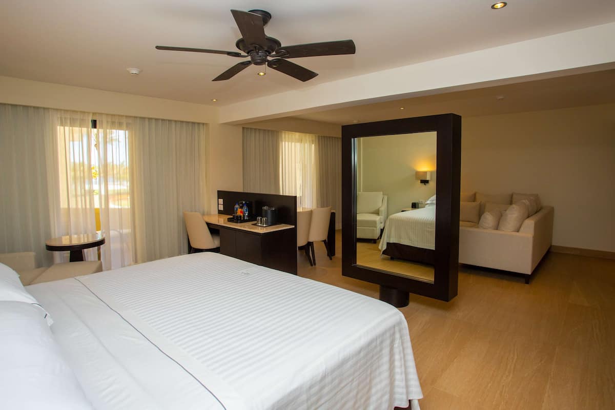 A hotel room with a bed, a desk, a chair, a sofa, a ceiling fan, and a large mirror.