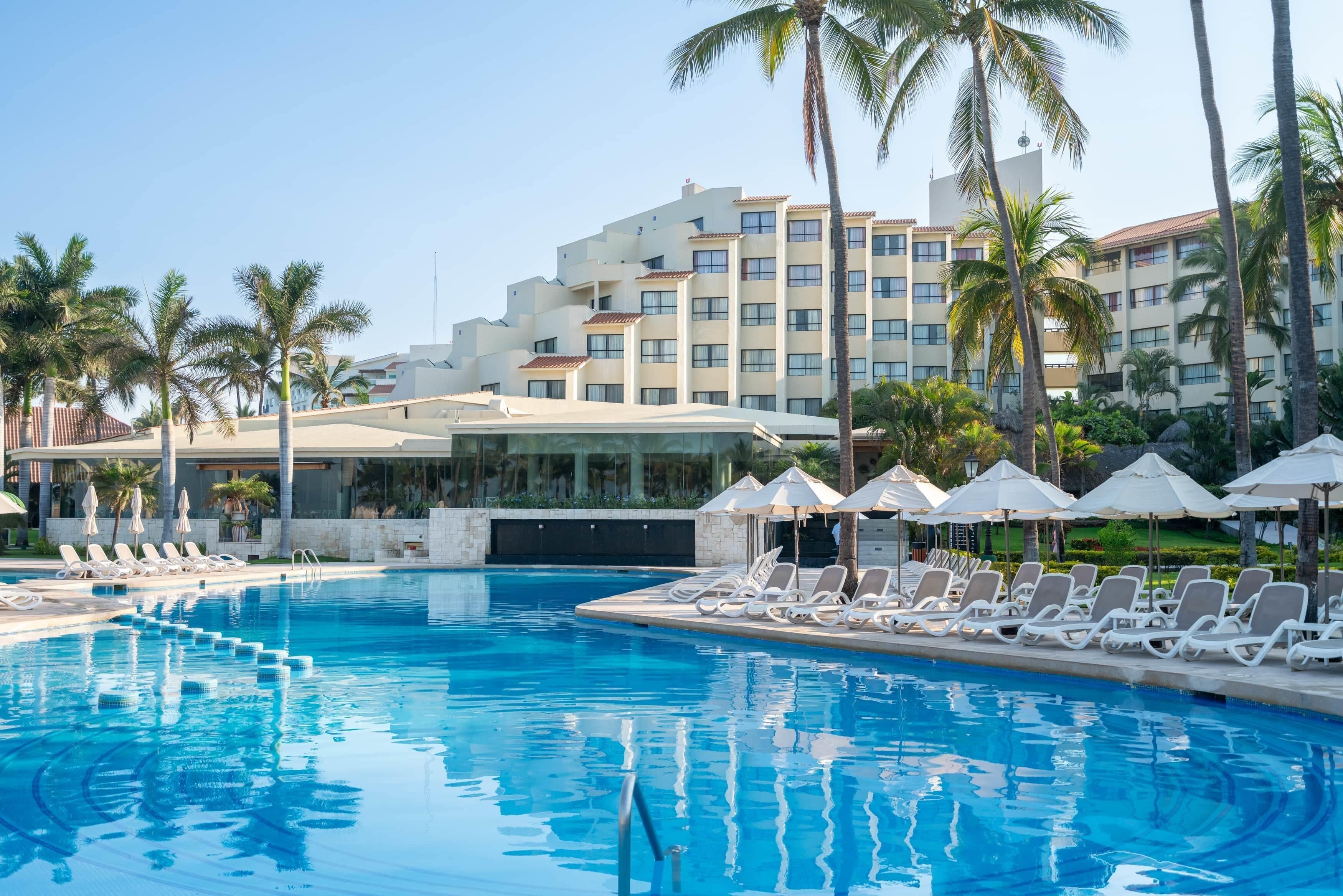 A swimming pool with lounge chairs and umbrellas, surrounded by palm trees and a hotel building.