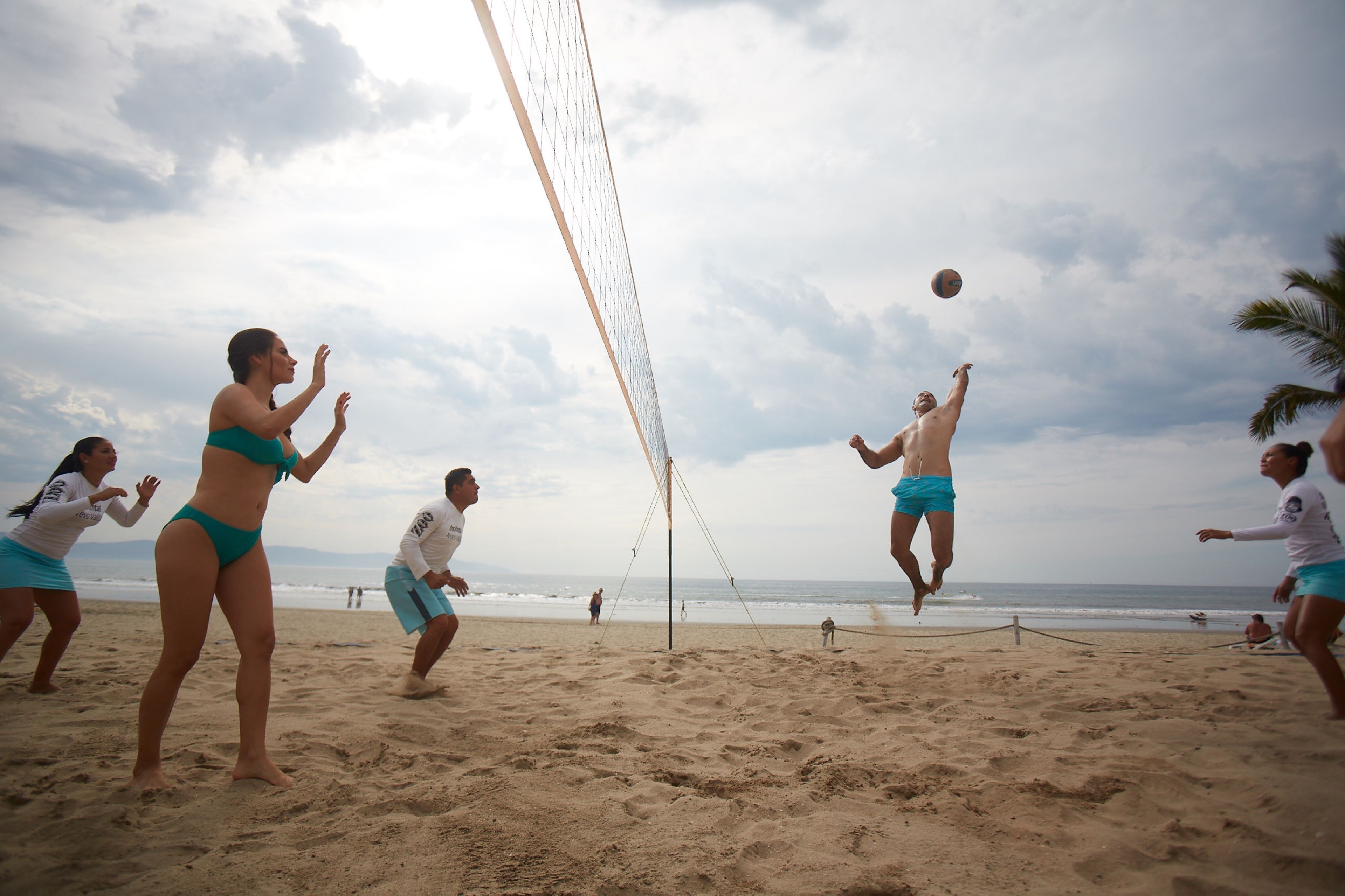 A beach volleyball game with players in bikinis and shorts, a volleyball net, and the ocean in the background.