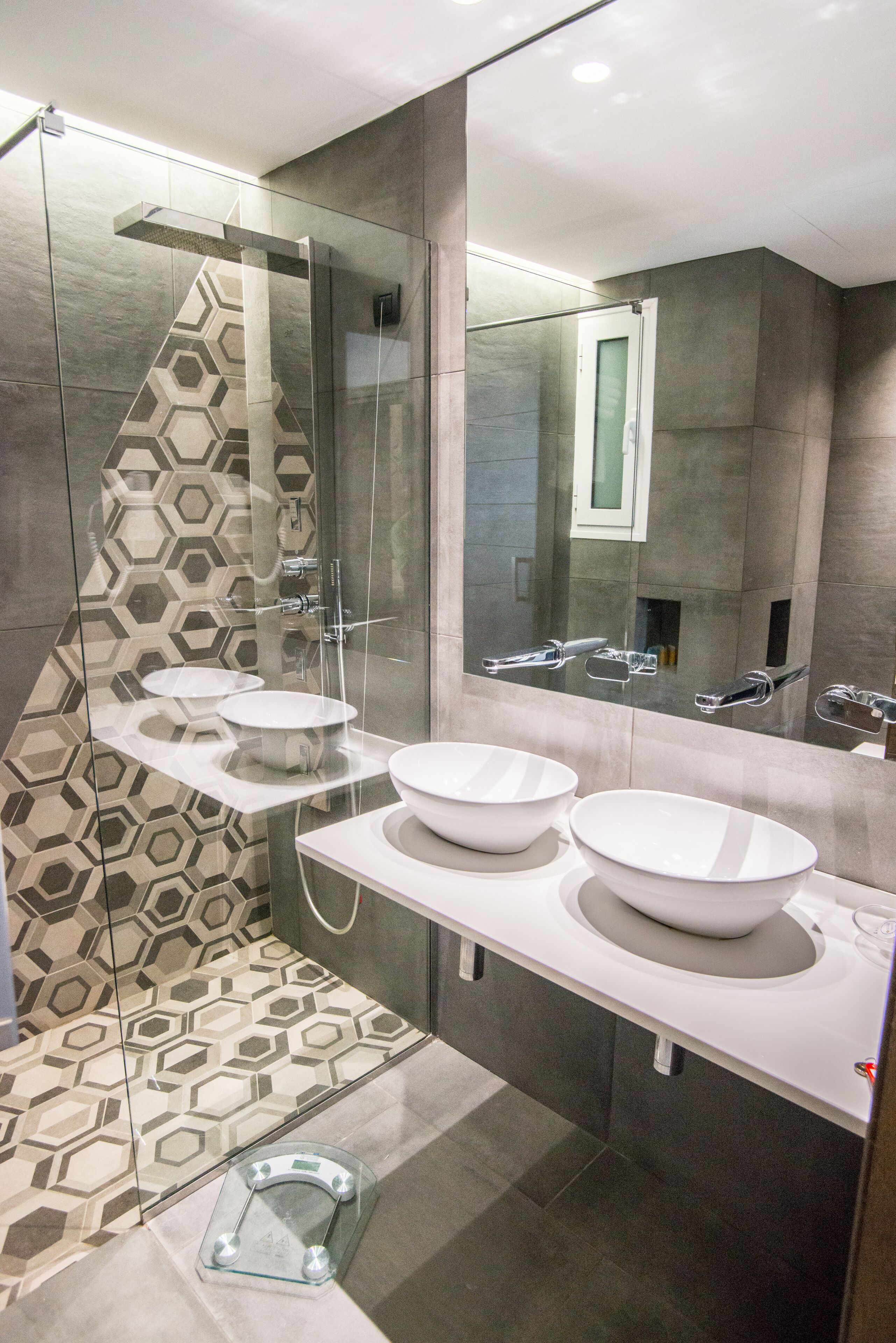 A modern bathroom with two white sinks, a glass shower enclosure, and a hexagonal patterned tile wall.