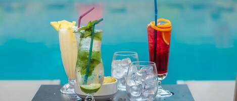 Three cocktails with garnishes and ice cubes on a table by a pool.