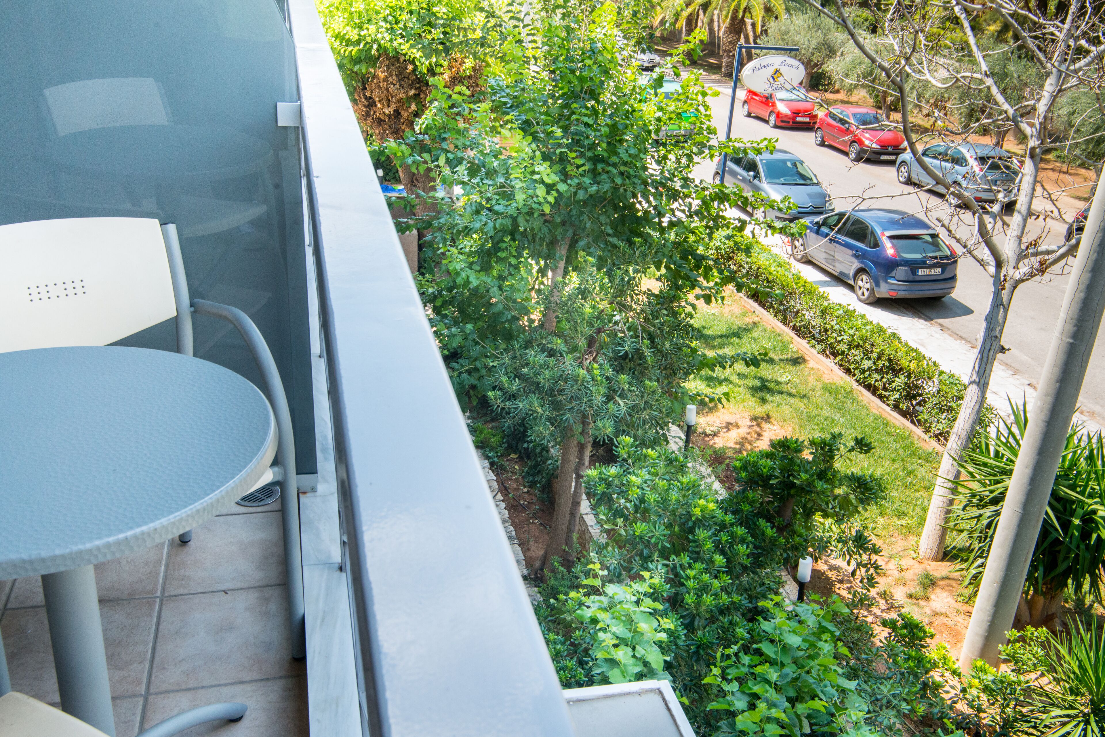 A balcony with a table and chairs, overlooking a street with parked cars and greenery.