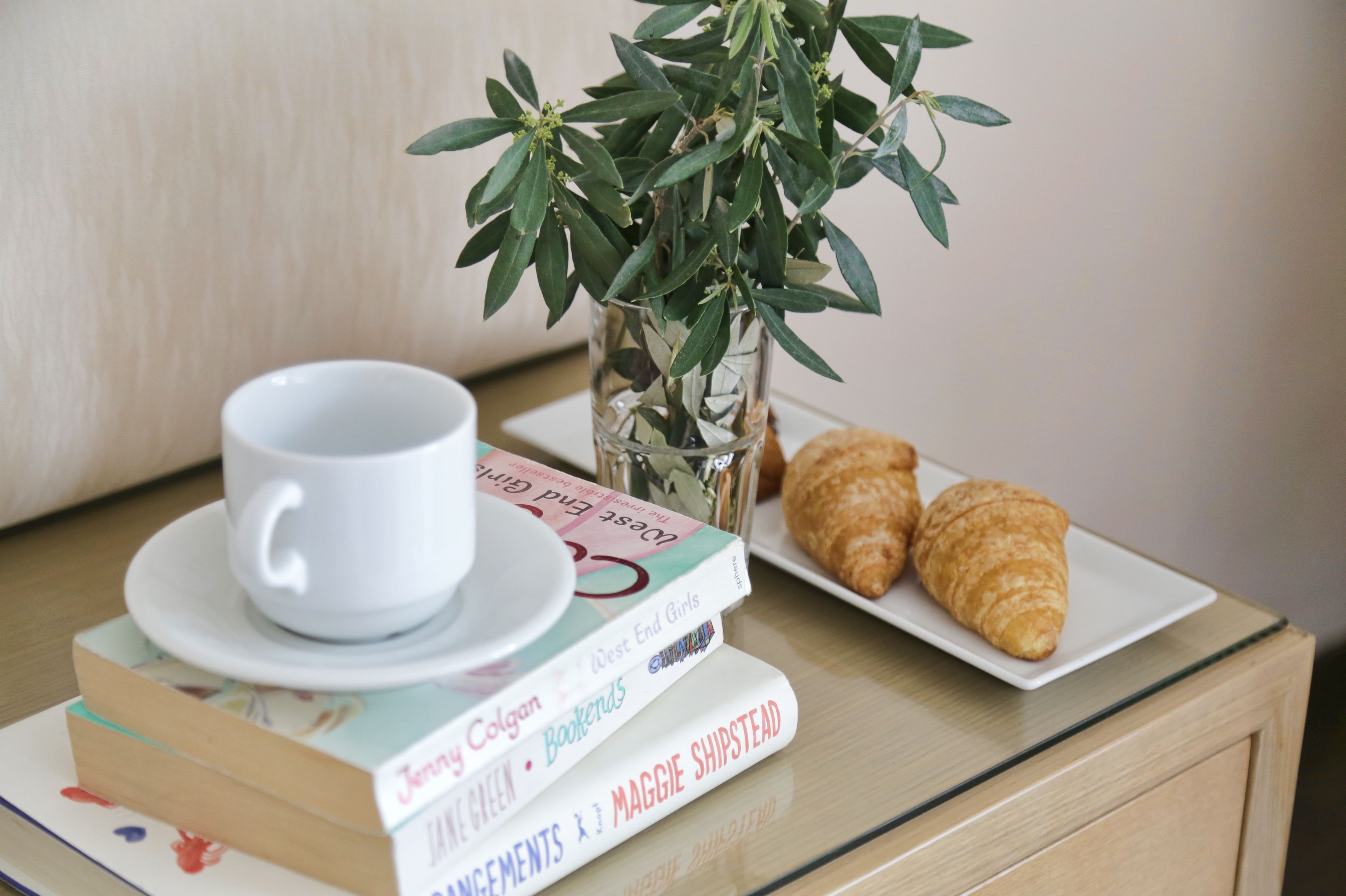 A white cup on a saucer, a glass vase with green leaves, and two croissants on a white plate, with books stacked beside them.