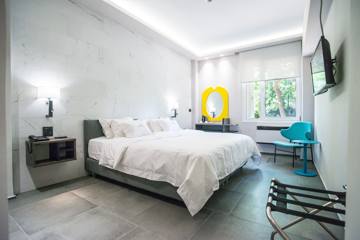 A modern bedroom with a bed, bedside tables, a chair, a TV, and a window with a view of trees.
