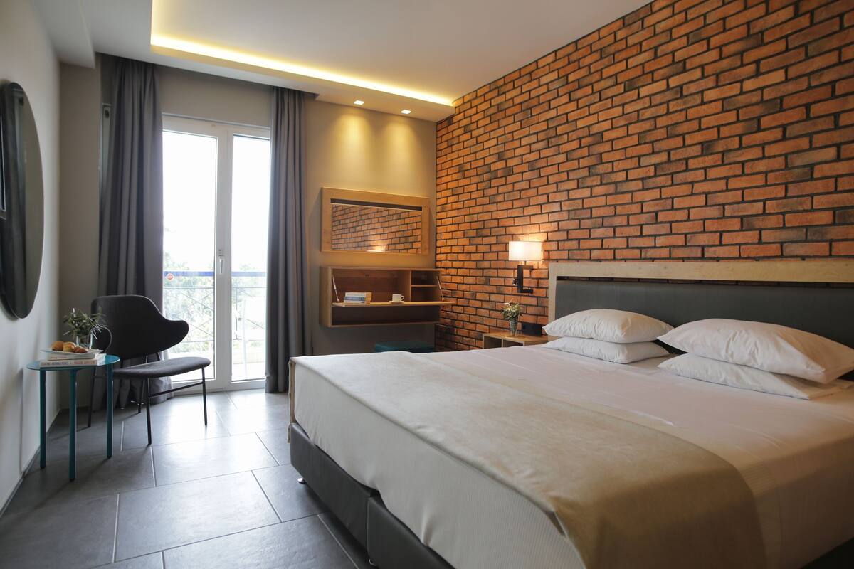 Una habitación de hotel moderna con una cama grande, una mesita pequeña, una silla, una mesita de noche y una pared con diseño de ladrillo.