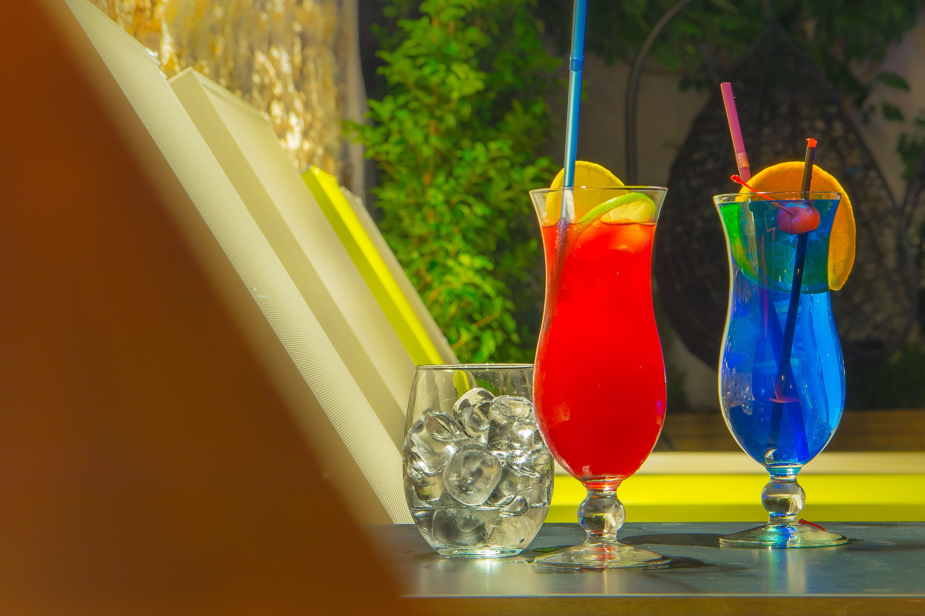 Two colorful cocktails with straws and lemon slices, ice cubes, and a glass of water.