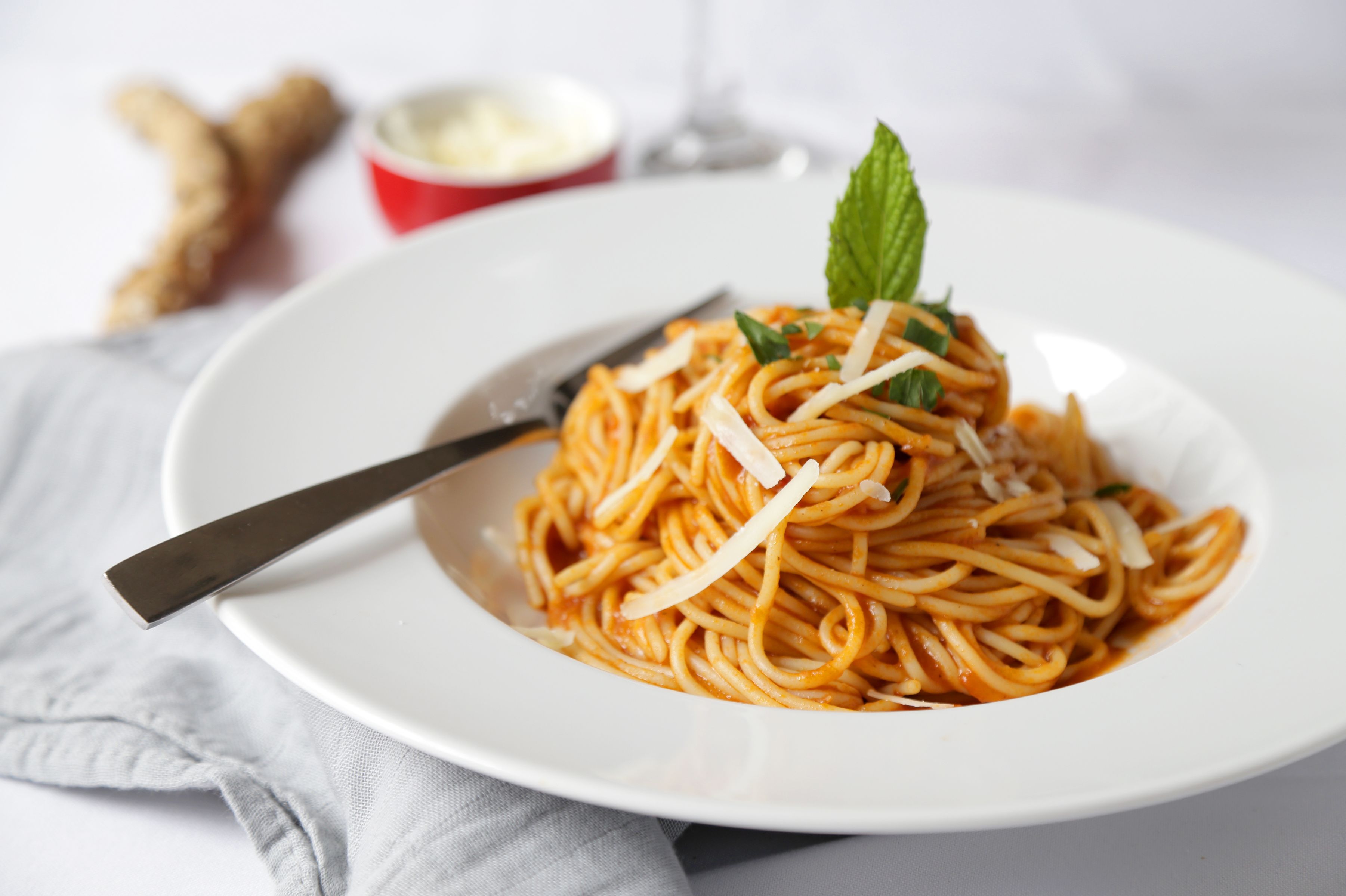 A plate of spaghetti with a mint leaf garnish, a fork, and a small bowl of sauce on a white plate.
