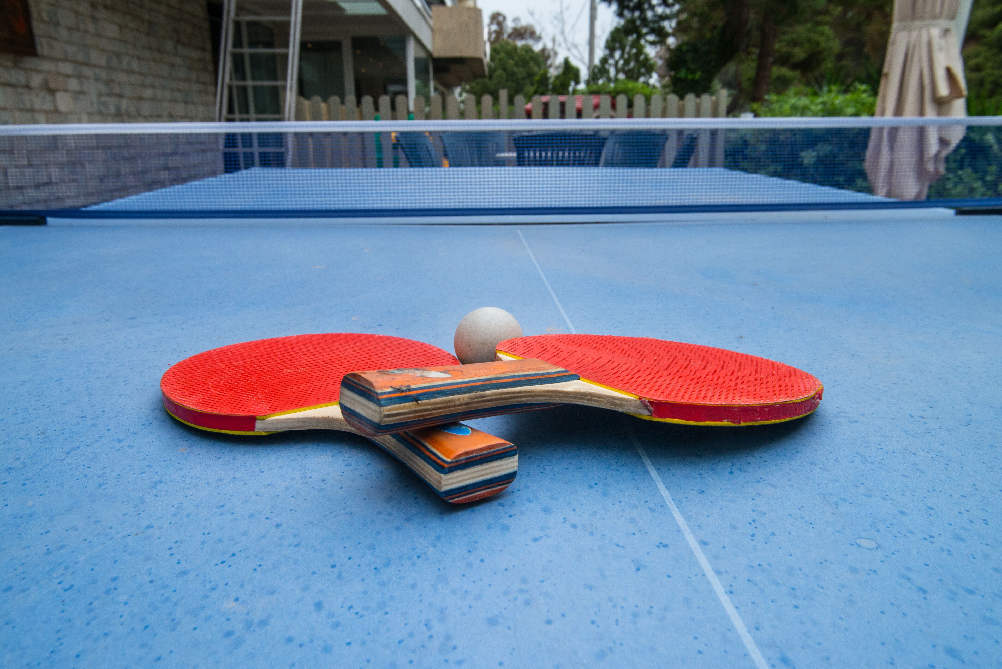 A ping pong table with paddles and a ball, set outdoors with a blue surface.