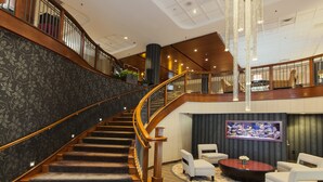 A grand staircase with a wooden railing leading to a second floor, a flat-screen TV mounted on the wall, and a seating area with a round coffee table.