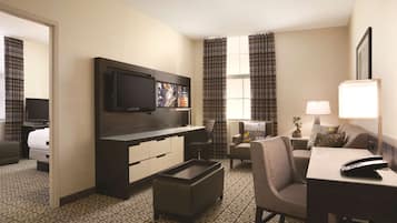 A hotel room with a flat-screen TV, a sofa, a dining table, a small coffee table, and a window with curtains.