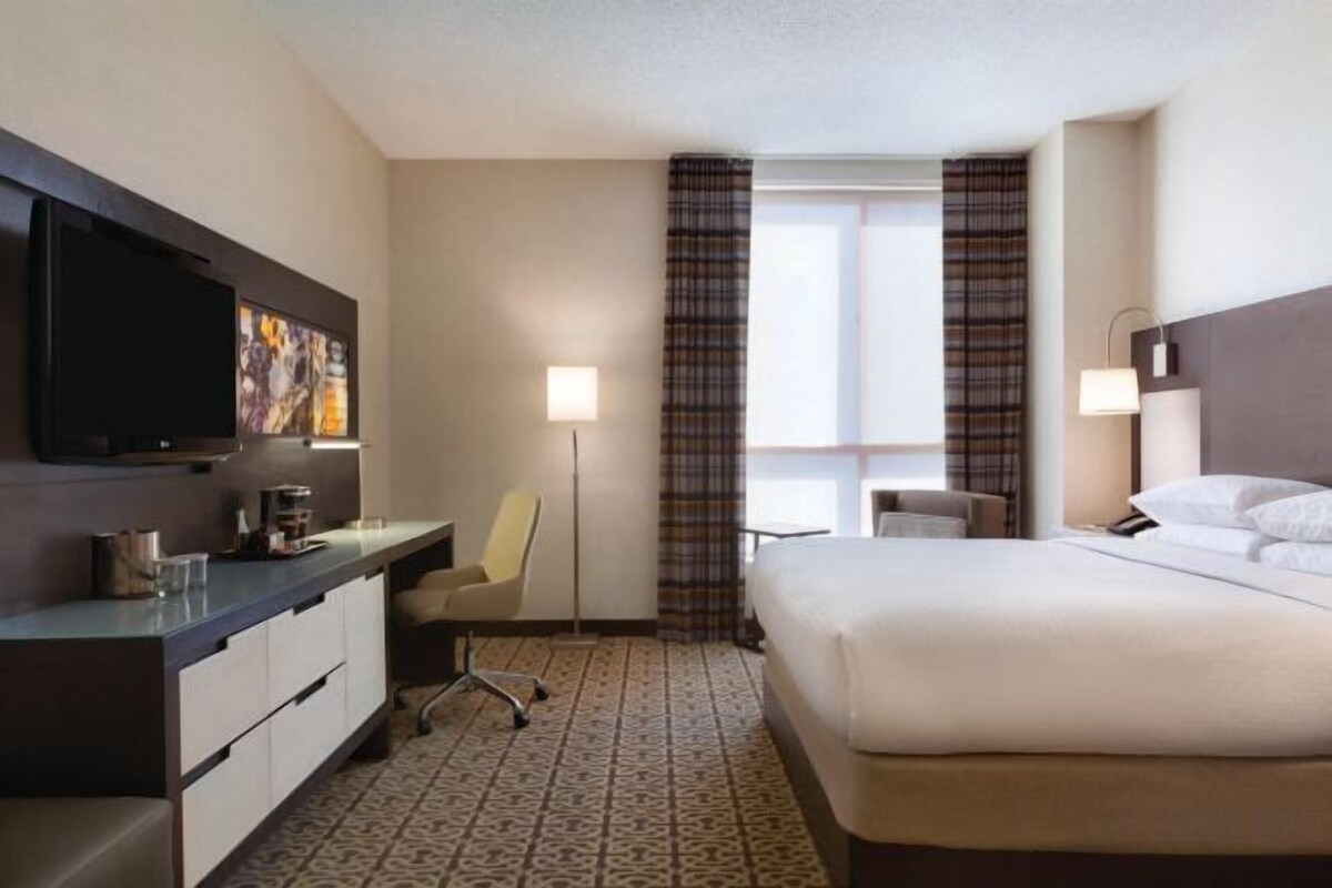 Une chambre d’hôtel comprenant un lit, un bureau avec une chaise, une télévision et une fenêtre avec des rideaux.