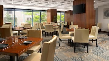 A spacious hotel dining area with multiple tables and chairs, large windows offering views of greenery, and a flat-screen TV mounted on a wooden panel.