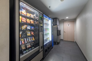 Un couloir avec des distributeurs automatiques proposant des collations et des boissons, une porte et un sol carrelé.