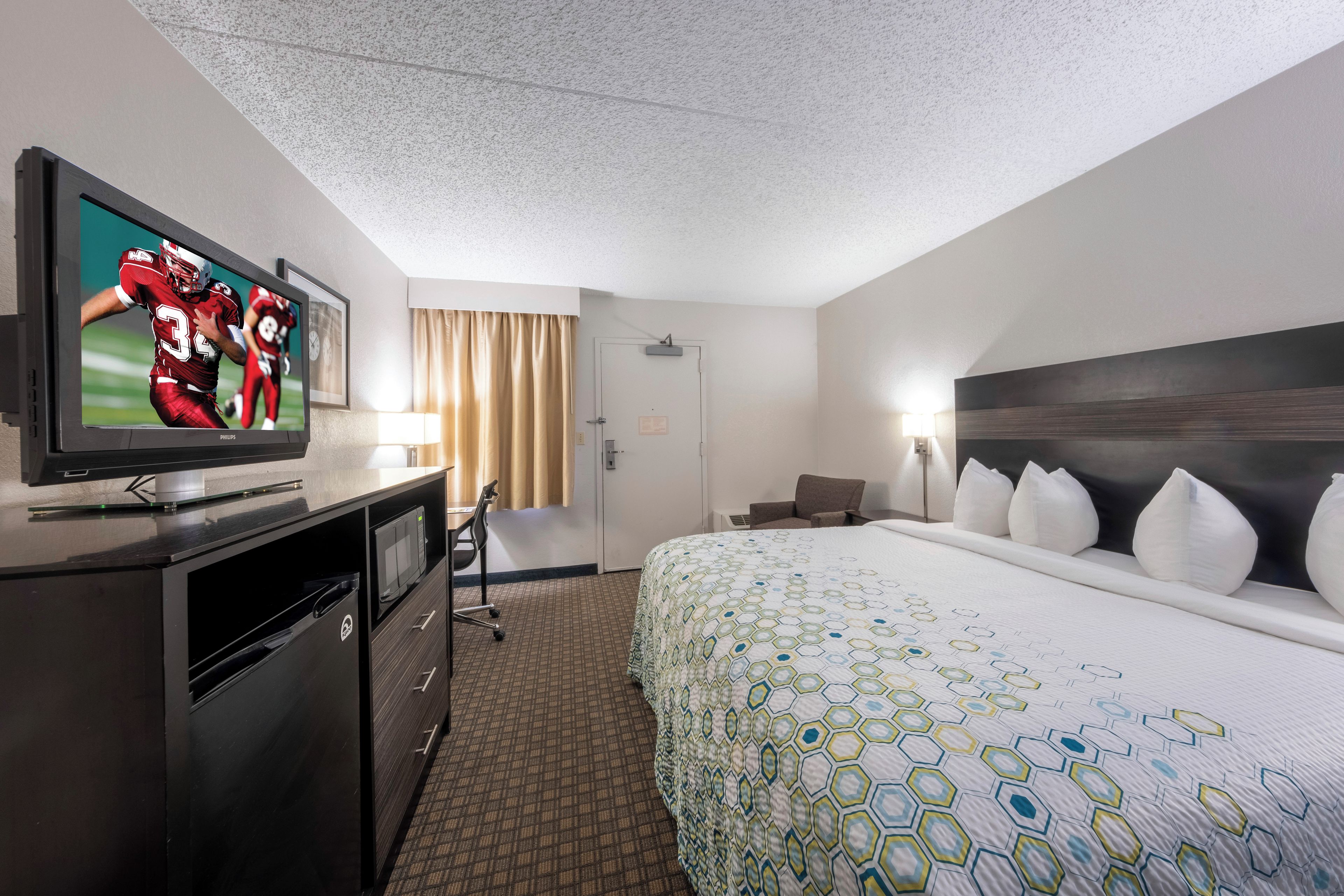 Una habitación de hotel con una televisión sobre un escritorio, una cama con colcha estampada, un escritorio con una silla y una puerta.