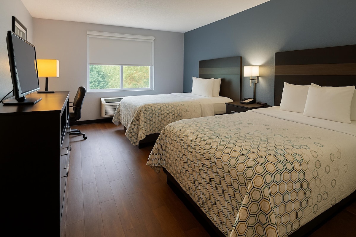 Une chambre d’hôtel avec deux lits, un bureau avec un ordinateur, une chaise, une fenêtre et une table de chevet avec une lampe.