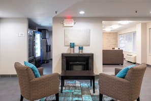 A modern interior with a fireplace, two wicker chairs, a table, a vending machine, and a reception desk. - HomeTown Inn by Red Roof East Syracuse (East Syracuse)