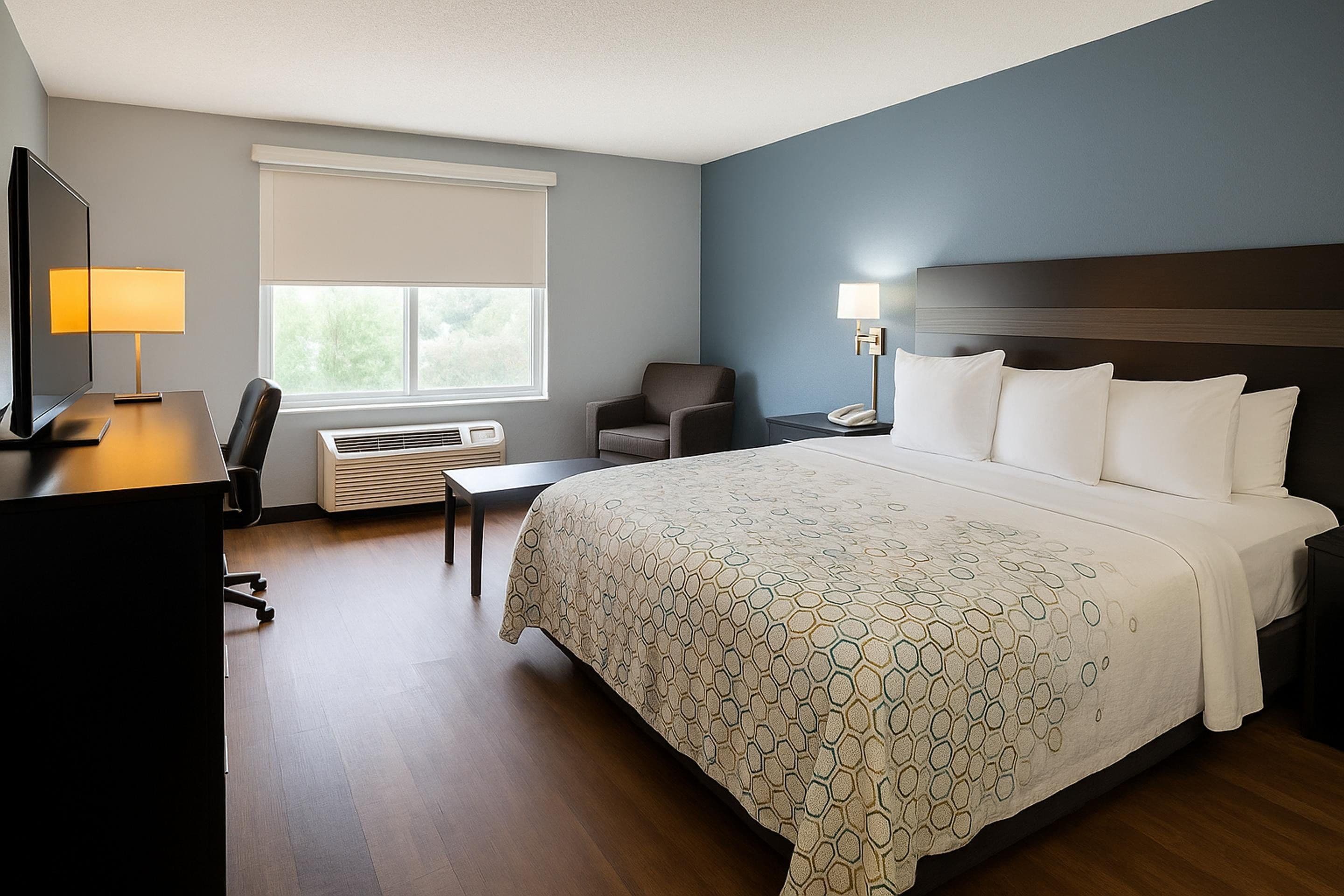 A hotel room with a large bed, a desk with a computer, a chair, a window with blinds, and an air conditioning unit.