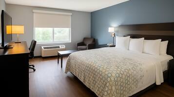 A hotel room with a large bed, a desk with a computer, a chair, a window with blinds, and an air conditioning unit.
