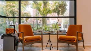 Two orange chairs with a small table between them, a suitcase, and a view of trees and buildings through large windows. - The Sunset Hotel West End (Vancouver)
