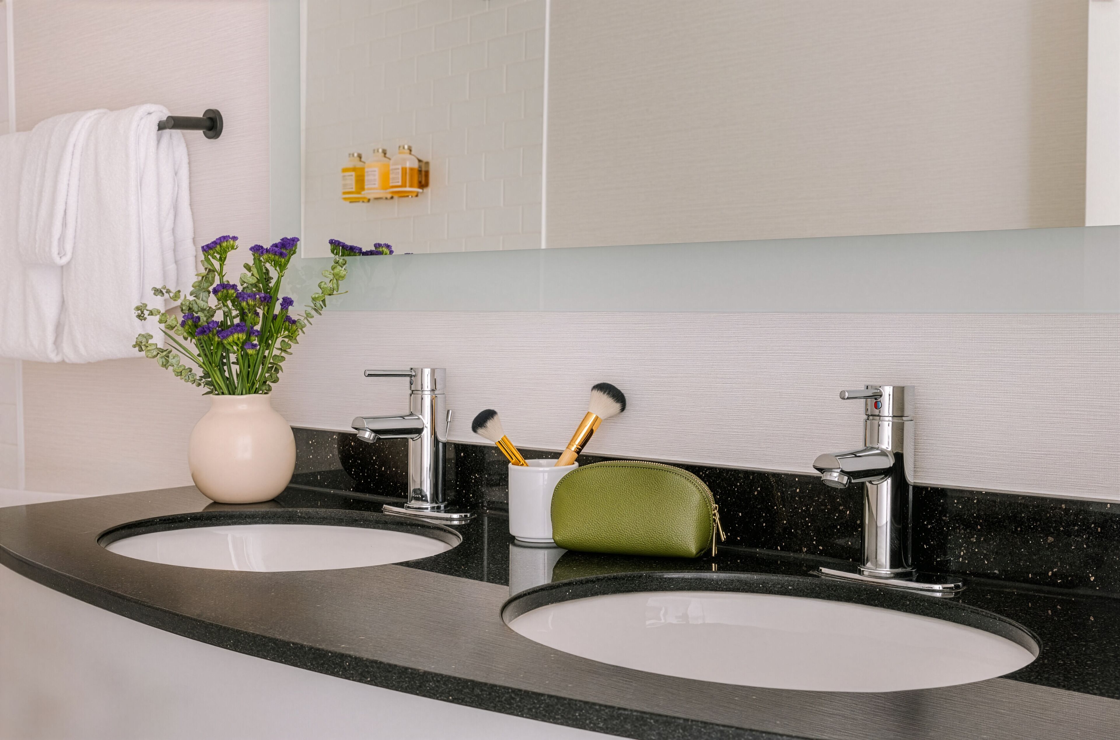 A bathroom with two sinks, a mirror, a towel rack, a vase with flowers, and a small shelf with toiletries.