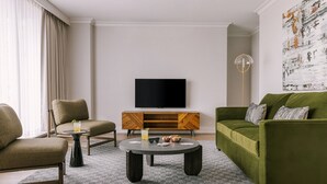 A modern living room with a green sofa, a beige armchair, and a round coffee table. A flat-screen TV is mounted on the wall above a wooden cabinet.