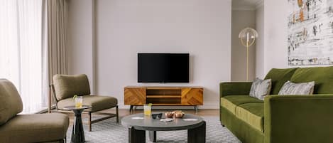 A modern living room with a green sofa, a beige armchair, and a round coffee table. A flat-screen TV is mounted on the wall above a wooden cabinet.