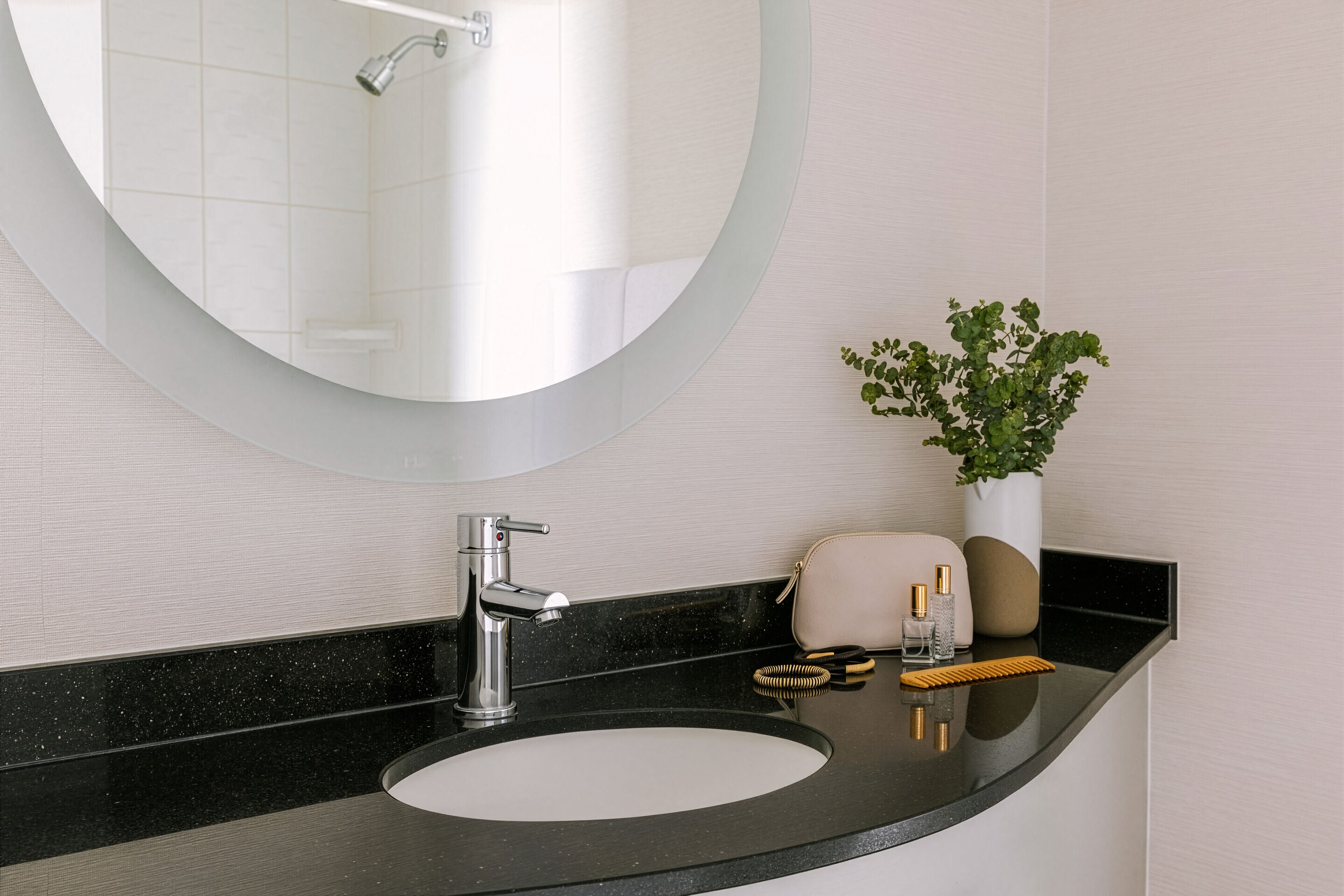A modern bathroom with a round mirror, a black countertop, a white sink, and a potted plant.