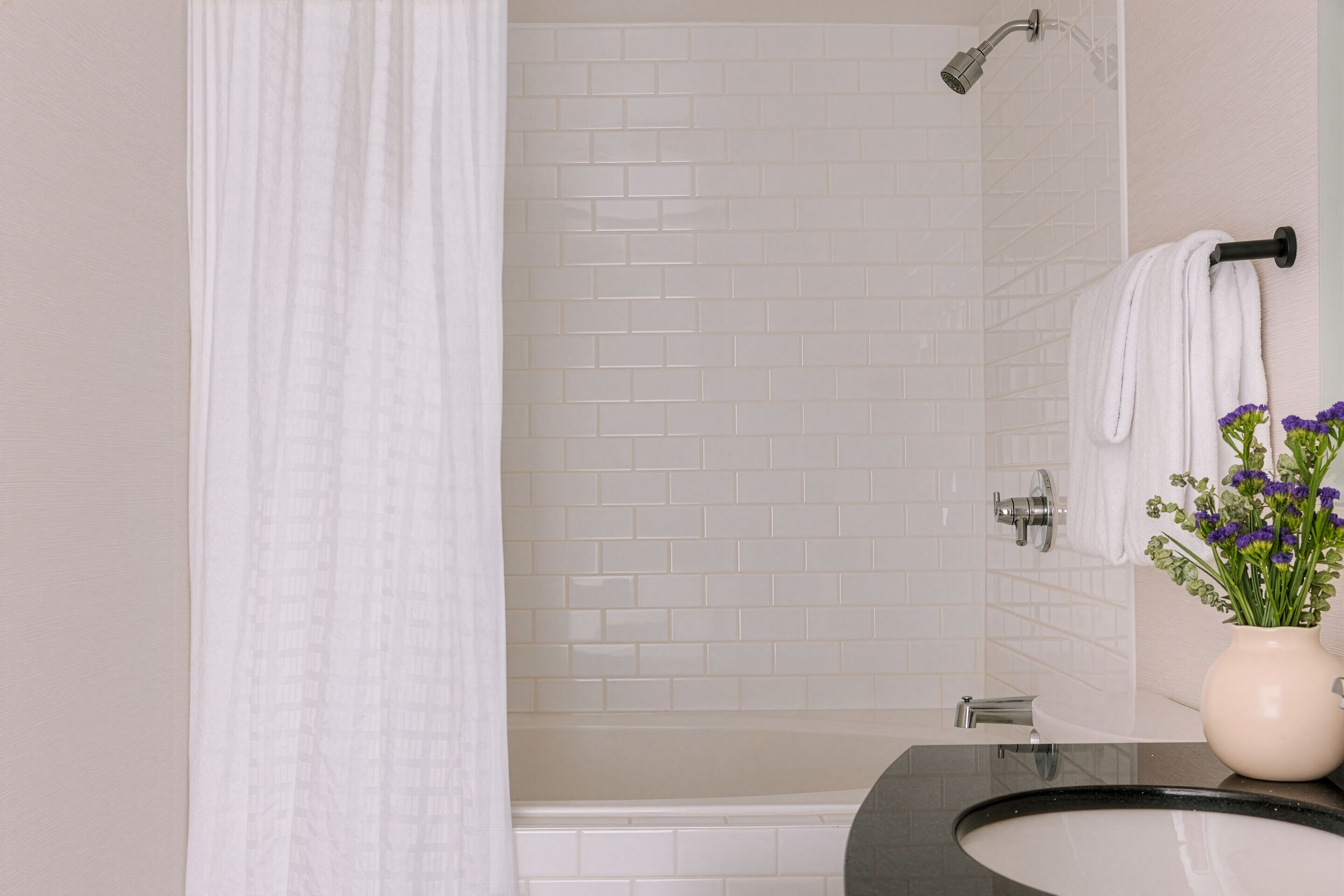 A bathroom with white tiled walls, a black sink, a shower with a handheld showerhead, and a vase with purple flowers on the counter.
