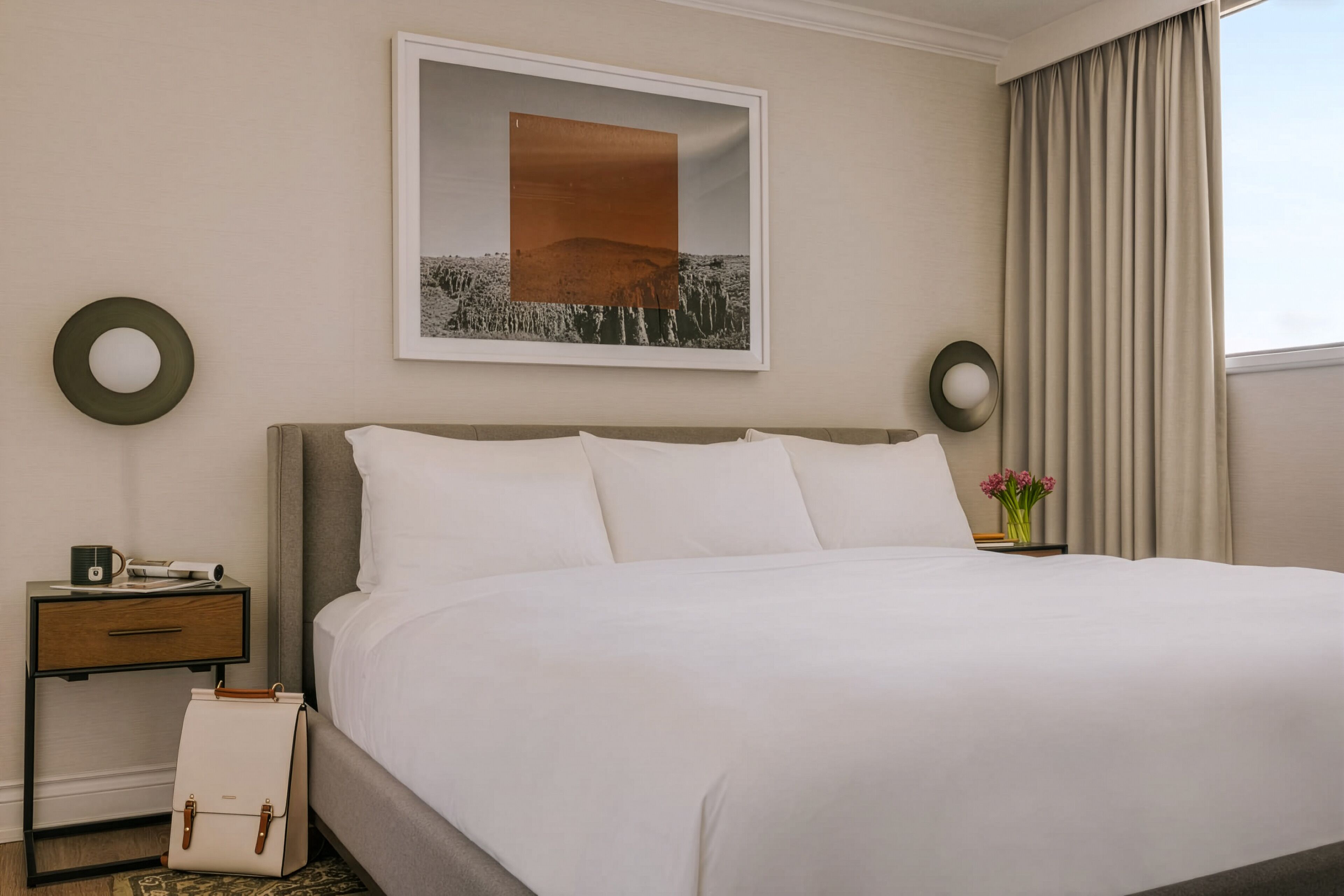 Habitación de hotel con una cama grande, una mesita de noche con lámpara, un cuadro enmarcado en la pared y una ventana con cortinas.