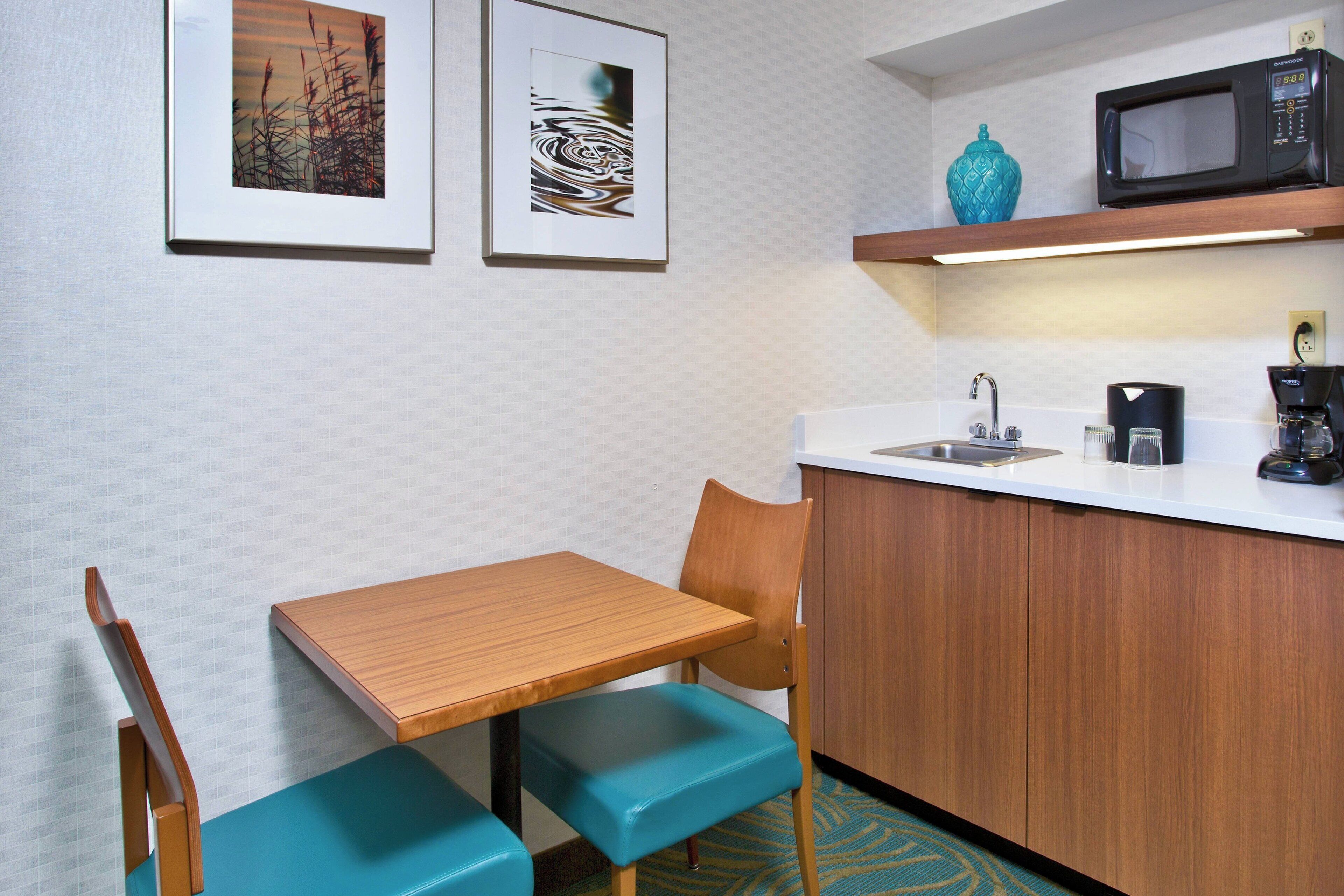 A compact kitchenette with a wooden table, teal chairs, a microwave, and a coffee maker.