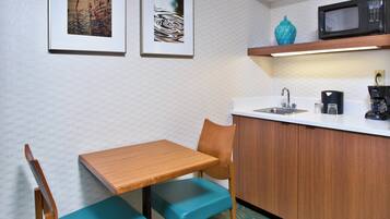 A compact kitchenette with a wooden table, teal chairs, a microwave, and a coffee maker.