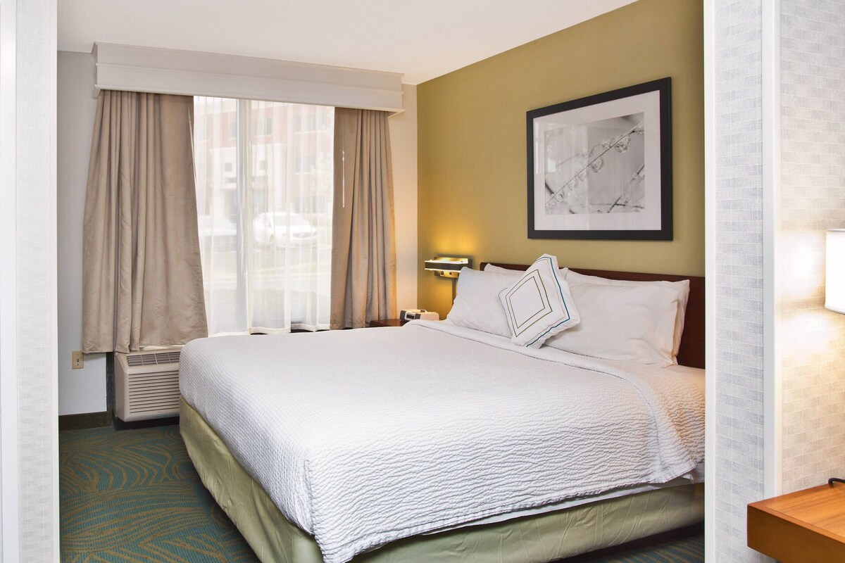 A hotel room with a large bed, beige curtains, a framed picture on the wall, and a nightstand with a lamp.