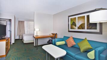 Habitación de hotel con cama, escritorio, silla, televisor, sofá con cojines y un cuadro en la pared.