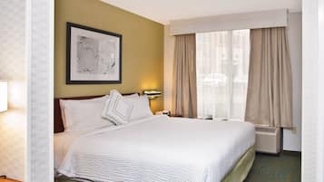 A hotel room with a large bed, a framed picture on the wall, a window with curtains, and a wall-mounted air conditioning unit.