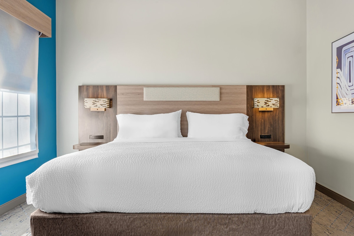 A modern hotel room with a large bed, wooden headboard, two bedside tables with lamps, and a large window.