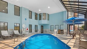 Piscina interior com paredes de vidro transparente, rodeada por espreguiçadeiras e uma mesa com guarda-sóis.