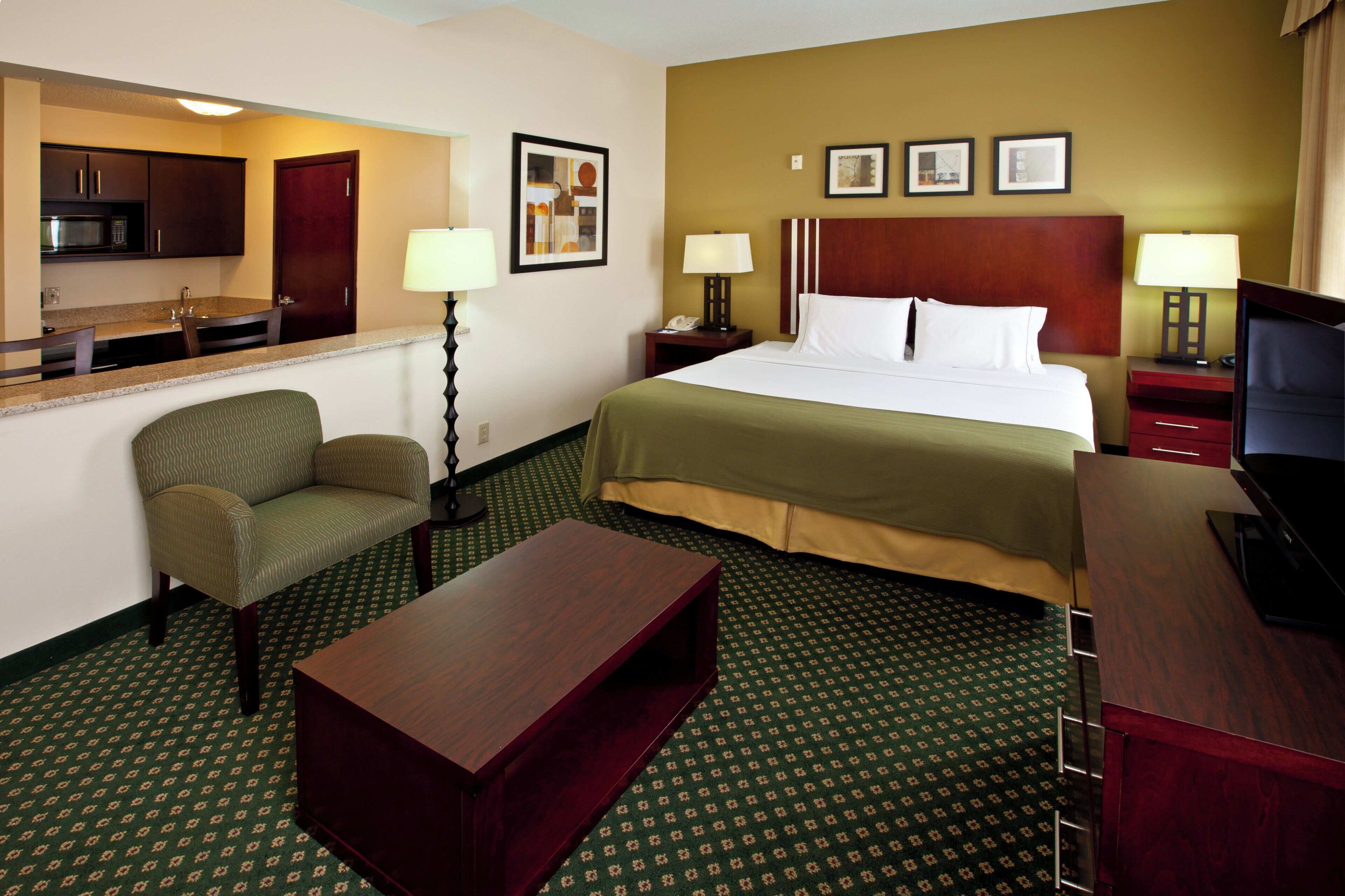 A hotel room with a bed, a desk, a chair, a small table, a lamp, a microwave, and a framed picture on the wall.