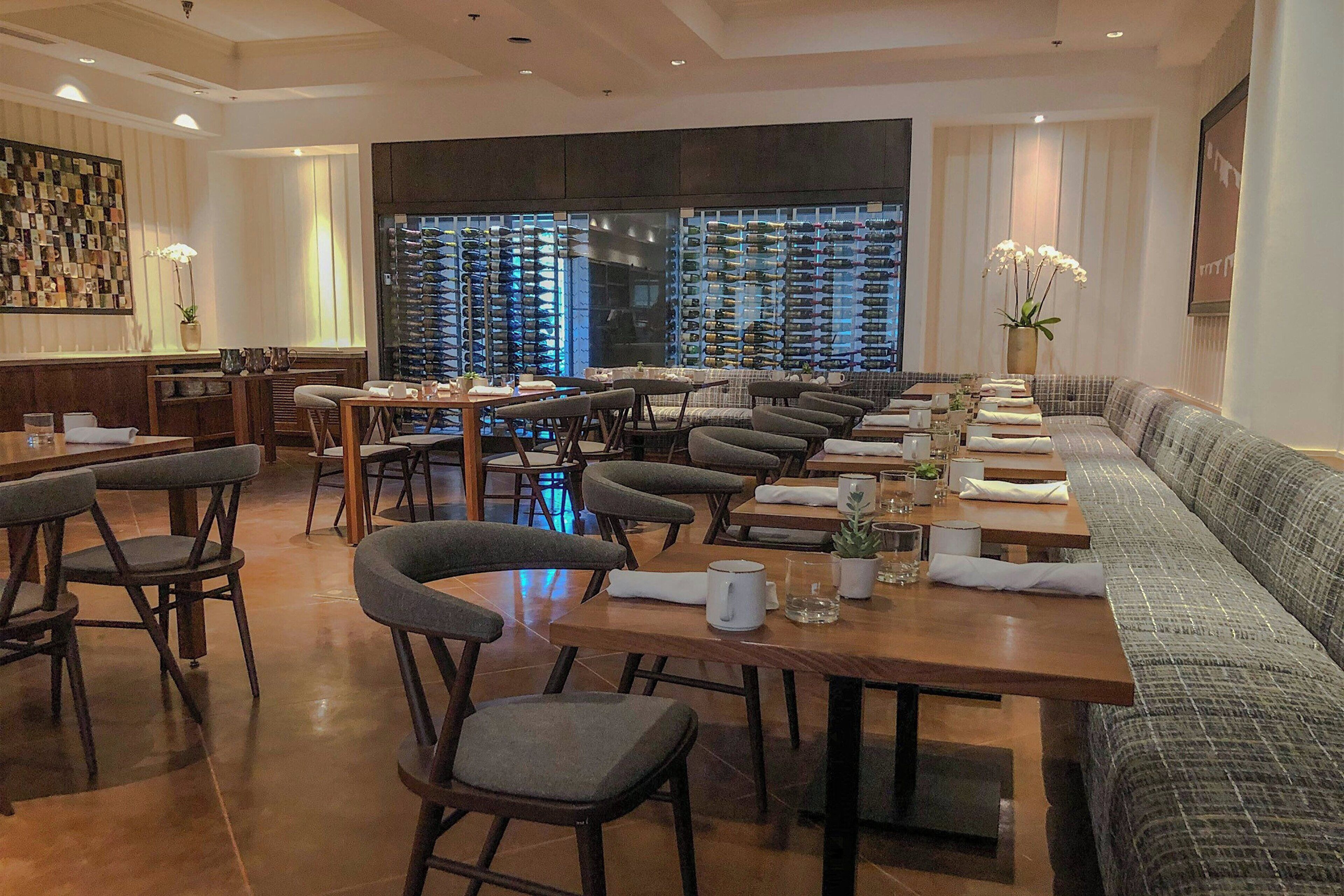 A restaurant with multiple dining tables and chairs, a long sofa, and a wall displaying numerous bottles.