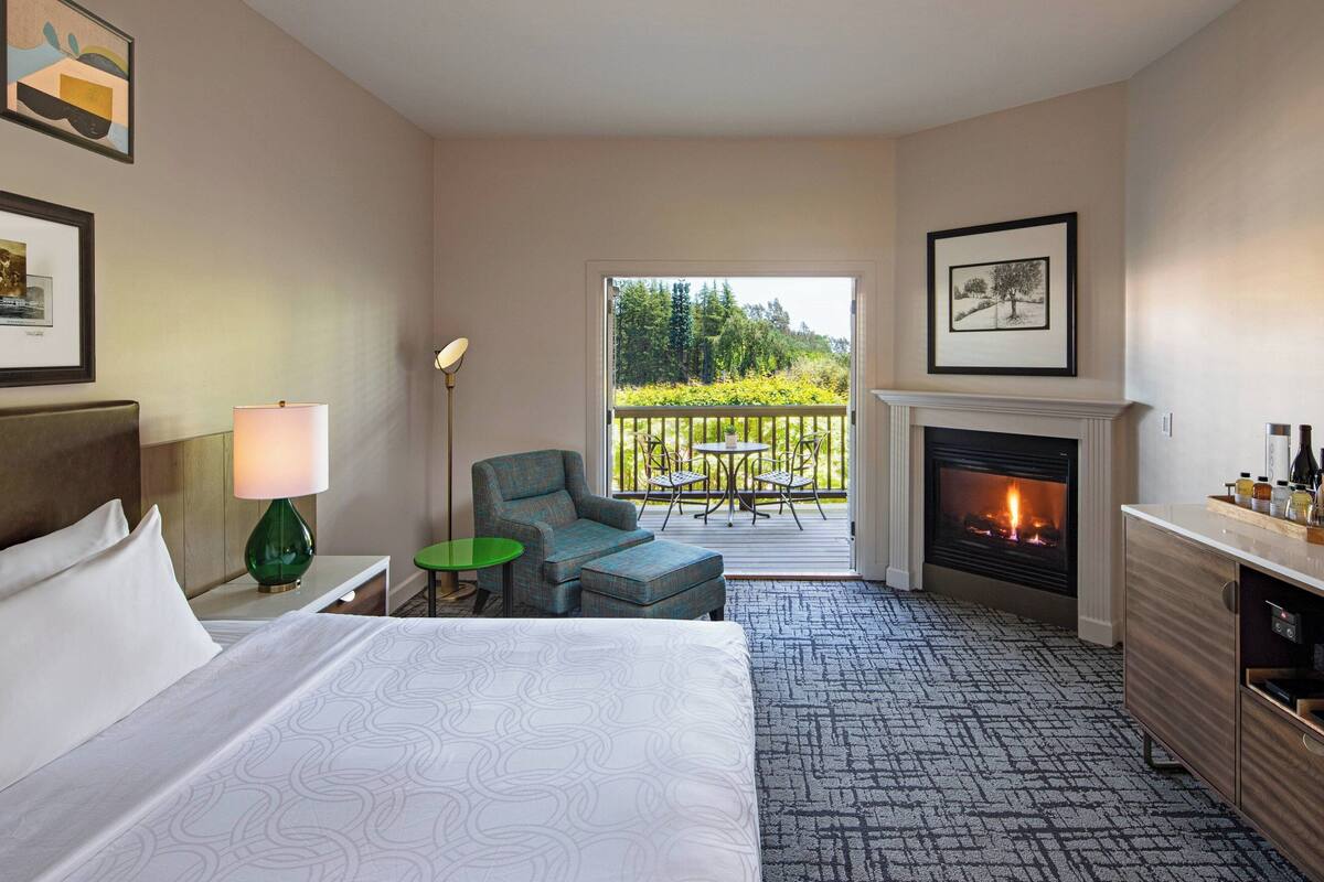 Hotelkamer met een bed, een groen nachtkastje, een lamp, een stoel, een open haard en een balkon met uitzicht op bomen.