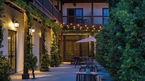 Un patio al aire libre con mesas y sillas, rodeado de follaje e iluminación con guirnaldas.