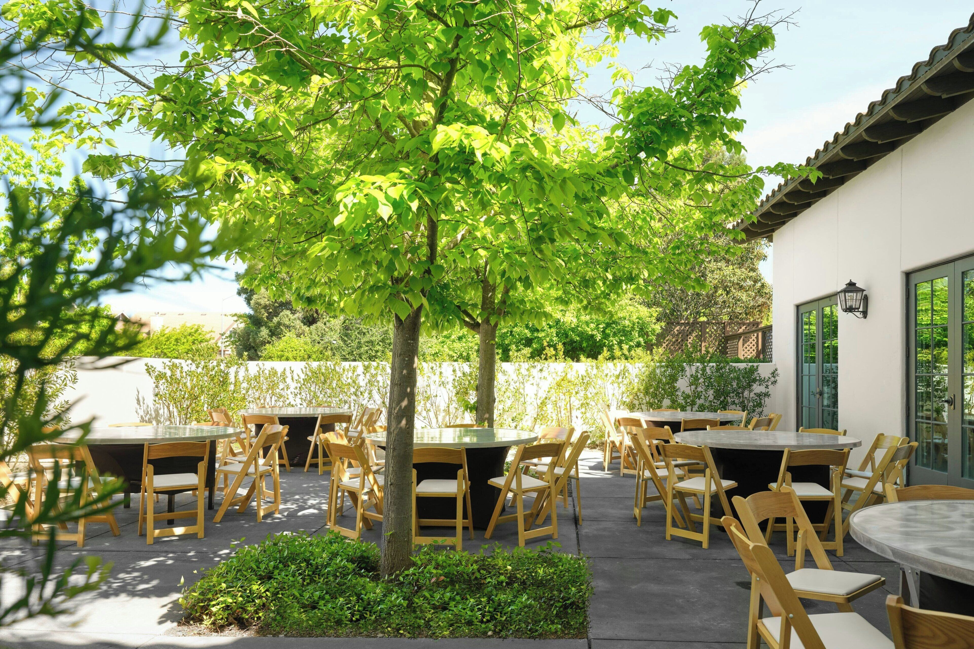 An outdoor patio with round tables and chairs, surrounded by greenery and a building with a tiled roof.