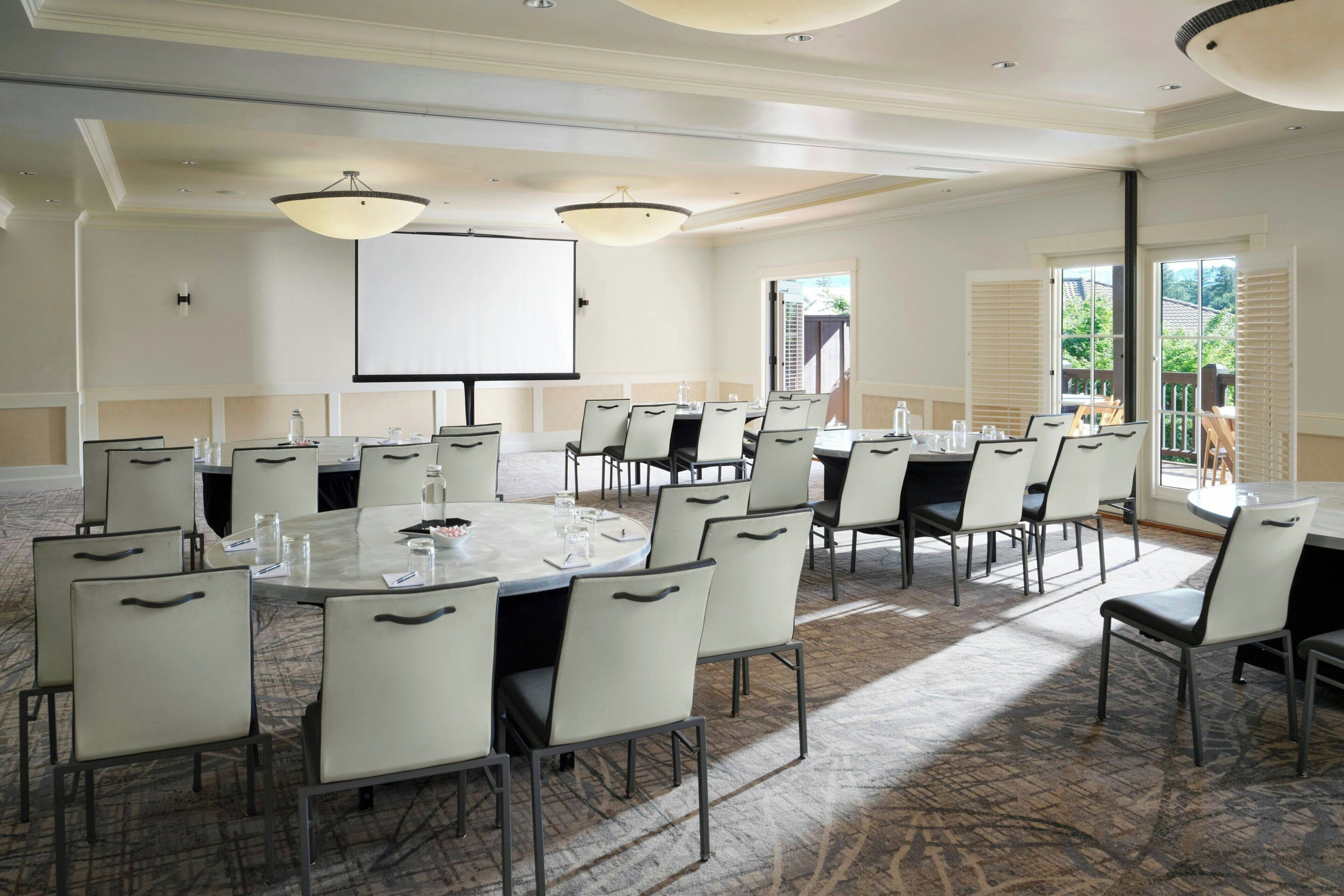 A conference room with a large table, multiple chairs, a projector screen, and a view of greenery outside.