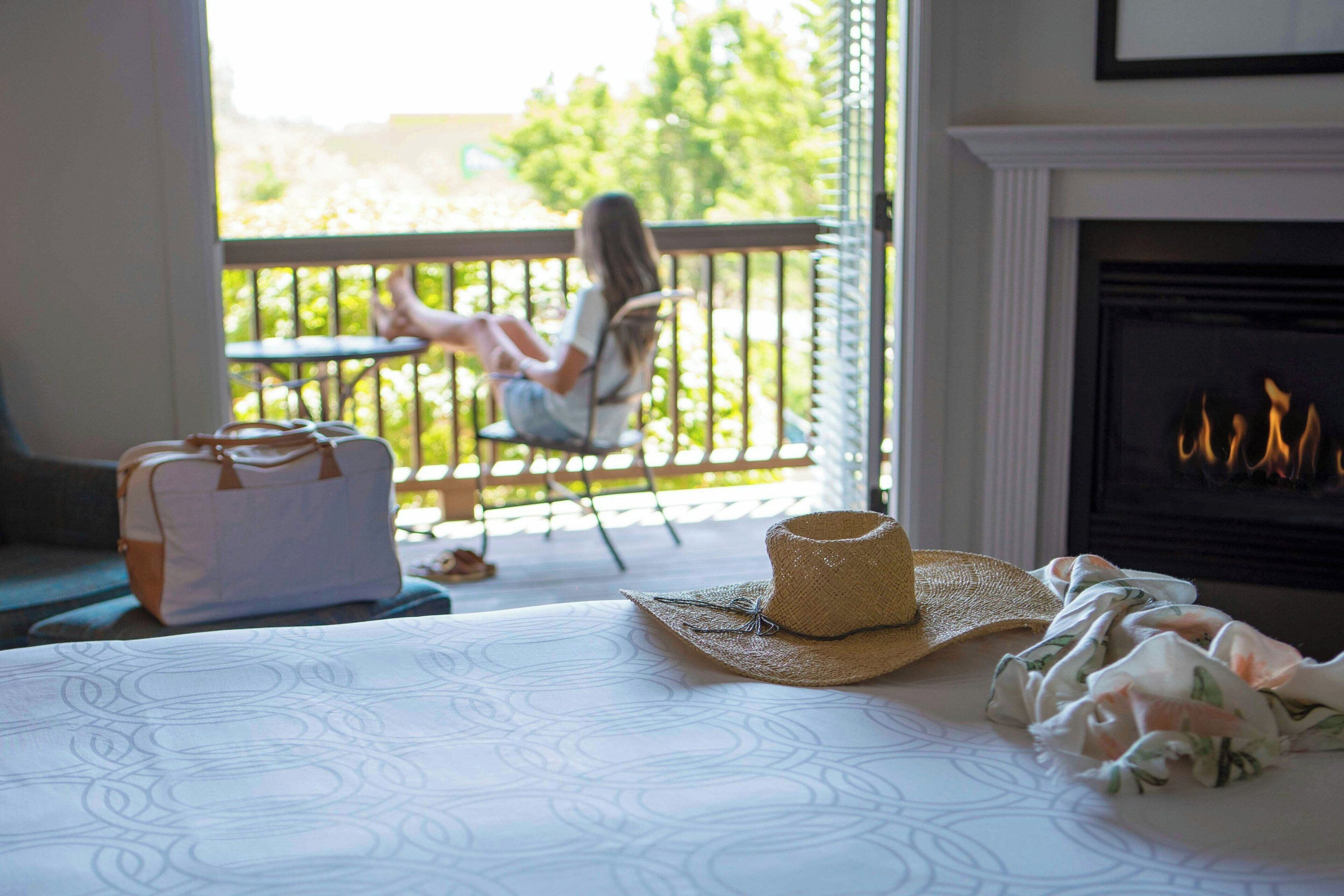 A room with a bed, a fireplace, a balcony with a person sitting, and a suitcase on the bed.