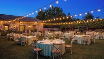 An outdoor banquet hall with round tables set for dining, string lights overhead, and a stone building in the background.