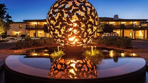 A spherical sculpture reflecting in a fountain, surrounded by a landscaped area with a building in the background.