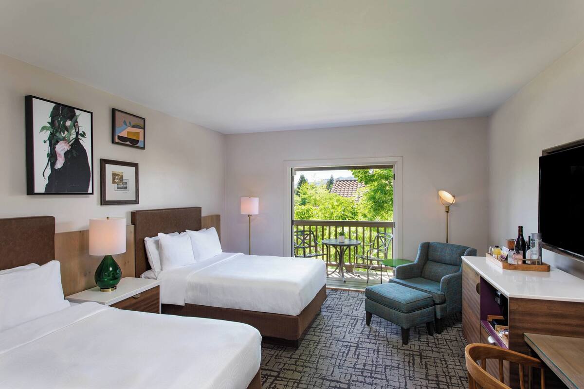 Een hotelkamer met twee bedden, een bureau, een stoel, een televisie en een balkon met uitzicht op groen.