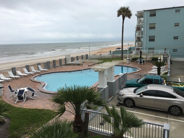 A pool area with lounge chairs, a car, and a building with a balcony overlooking the beach. - Arya Blu Inn & Suites (Ormond Beach)