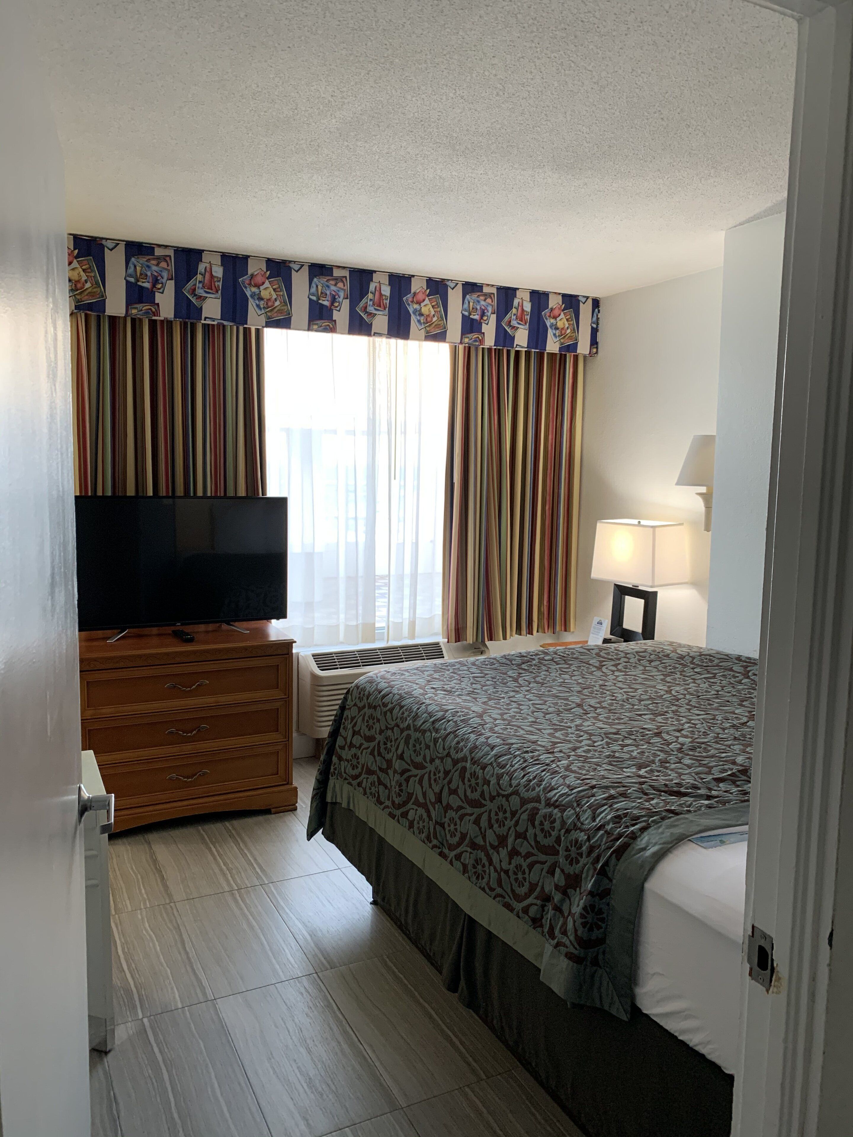 Una habitación de hotel con una cama, un televisor sobre un aparador de madera, una lámpara y cortinas.