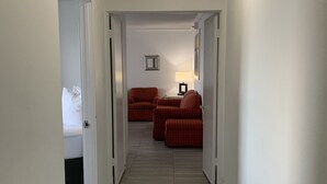 Passadís d'hotel amb un llit, dues cadires tapissades vermelles, una taula petita amb una làmpada i una llum al sostre.
