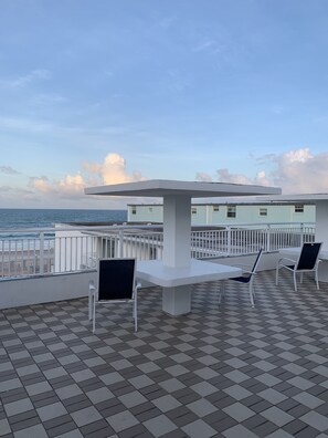 A terrace with a table and chairs, a view of the sea, and a building in the background. - Arya Blu Inn & Suites (Ormond Beach)